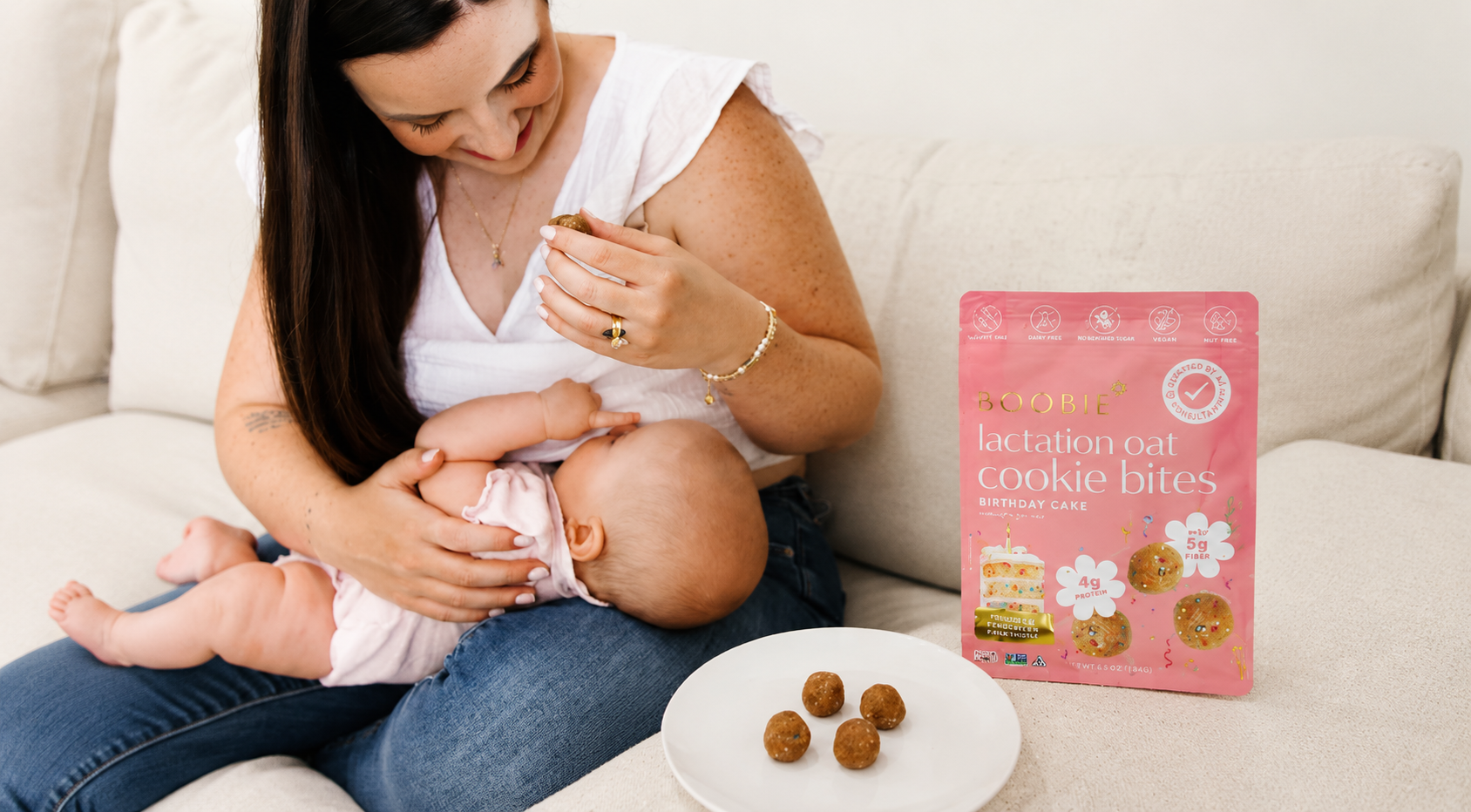 A breastfeeding mom snacking on BOOBIE Lactation Oat Cookie Bites while nursing her baby, with the Birthday Cake flavor bag displayed alongside a small plate of cookie bites.