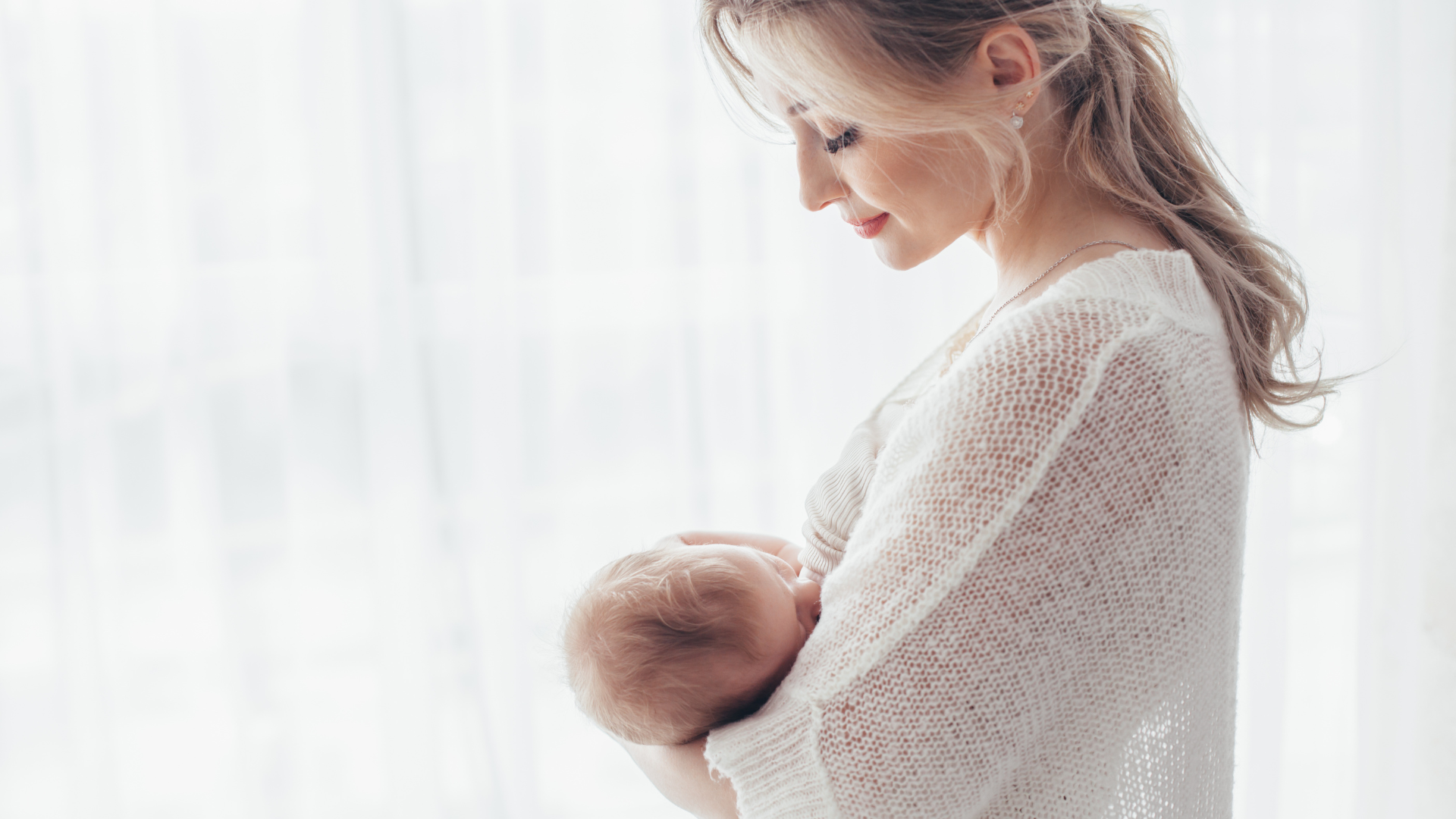 A mother in a cozy white sweater breastfeeding her newborn baby by a sunlit window, representing postpartum recovery and immune support.